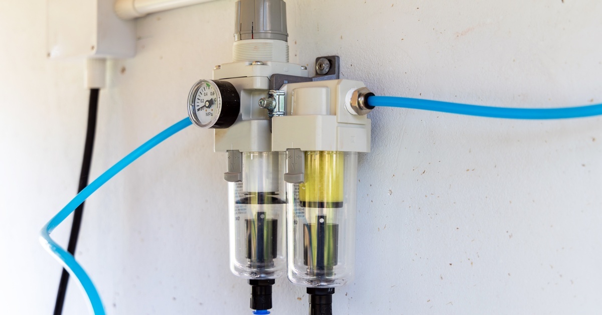 A white air pressure regulator unit with two blue tubes and a circular gauge is mounted to a white wall.