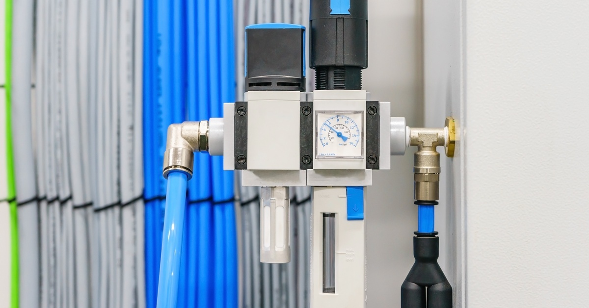 A white FRL unit with two blue tubes and a square check valve is mounted on a white wall with wires in the background.