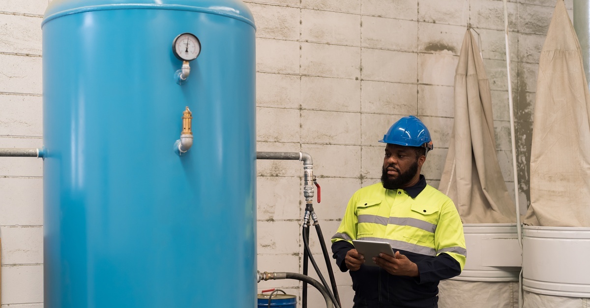 A large blue air receiver tank beside a worker in safety gear inspecting equipment in an industrial facility. A large blue air receiver tank beside a worker in safety gear inspecting equipment in an industrial facility.